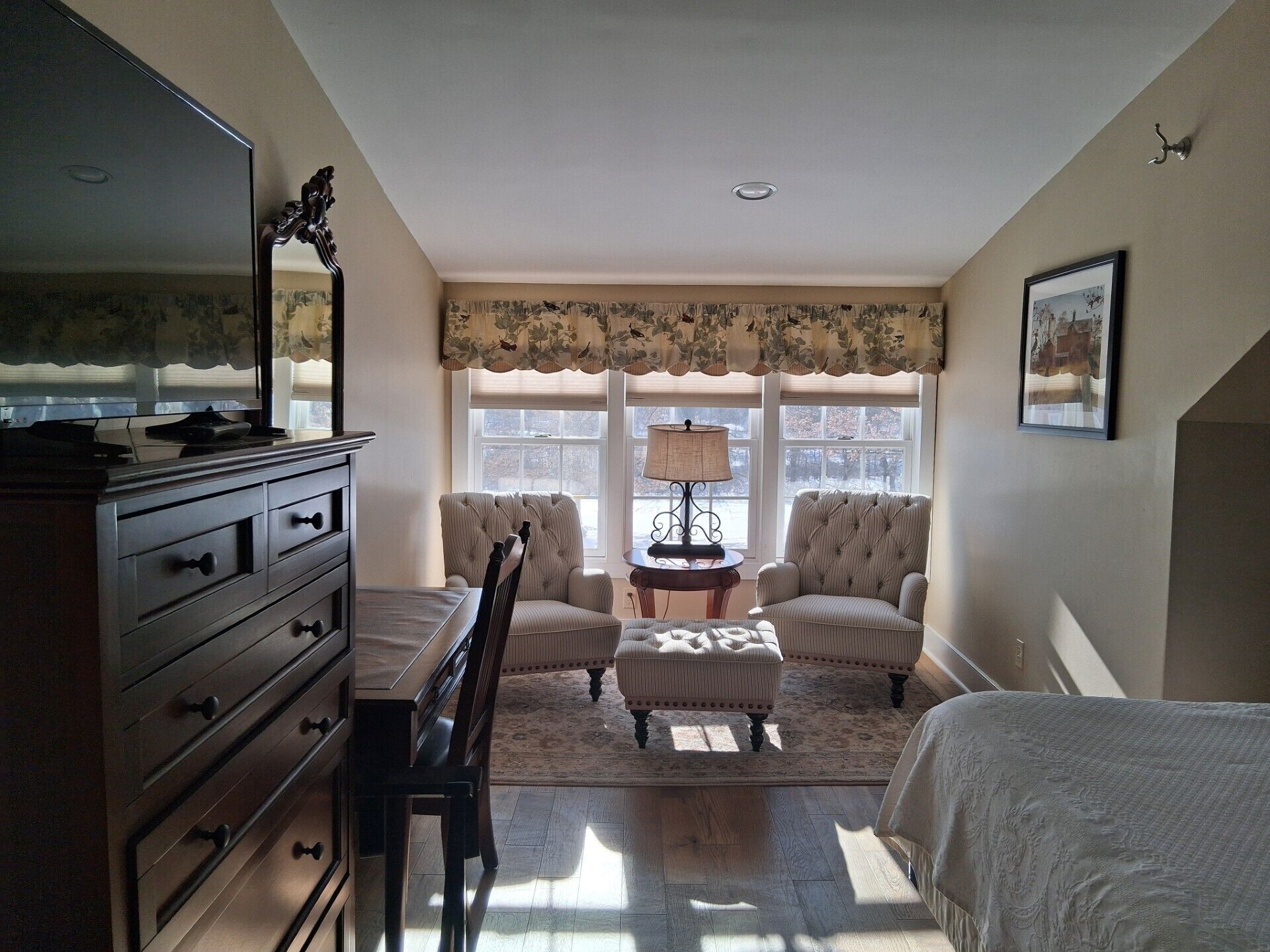 A bedroom with a bed, a dresser, a chair, a table with a lamp, and a window with curtains.