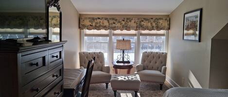 A bedroom with a bed, a dresser, a chair, a table with a lamp, and a window with curtains.
