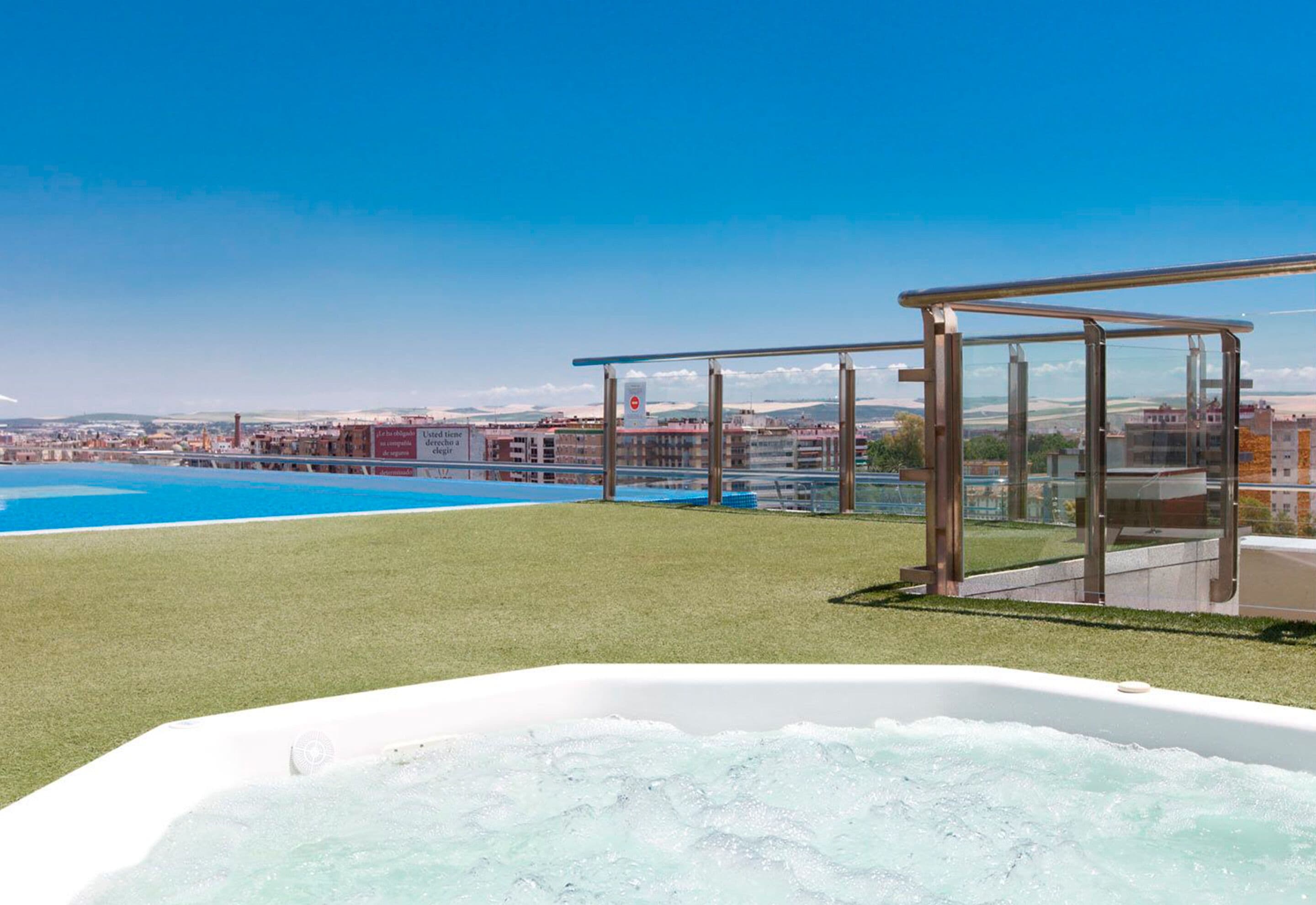A rooftop pool area with a hot tub, clear skies, and a view of the cityscape.