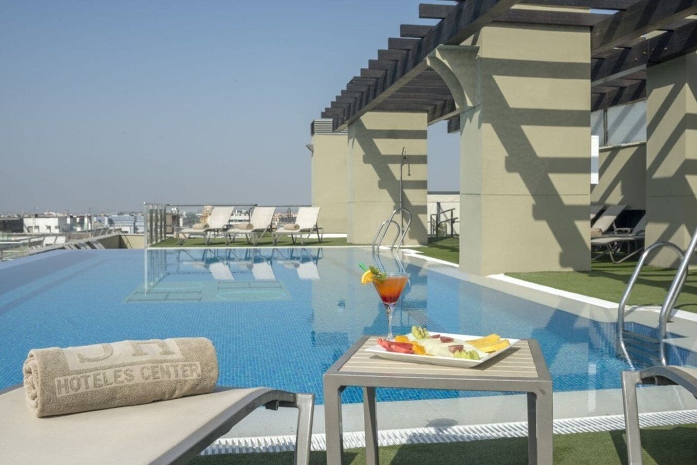 A rooftop pool with lounge chairs, a table with a drink and fruit, and a towel labeled "HOTELES CENTER".