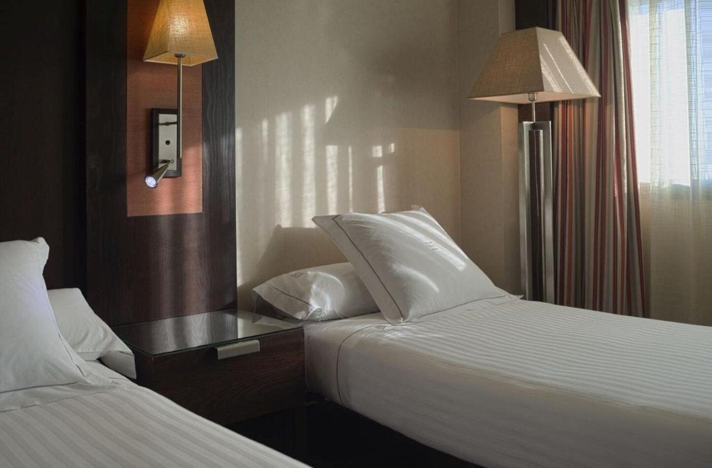 Une chambre d’hôtel avec deux lits, une table de chevet, une lampe, une horloge et une fenêtre avec des rideaux.