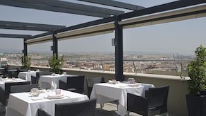 Un área de comedor al aire libre con mesas y sillas, con vista a un paisaje urbano.