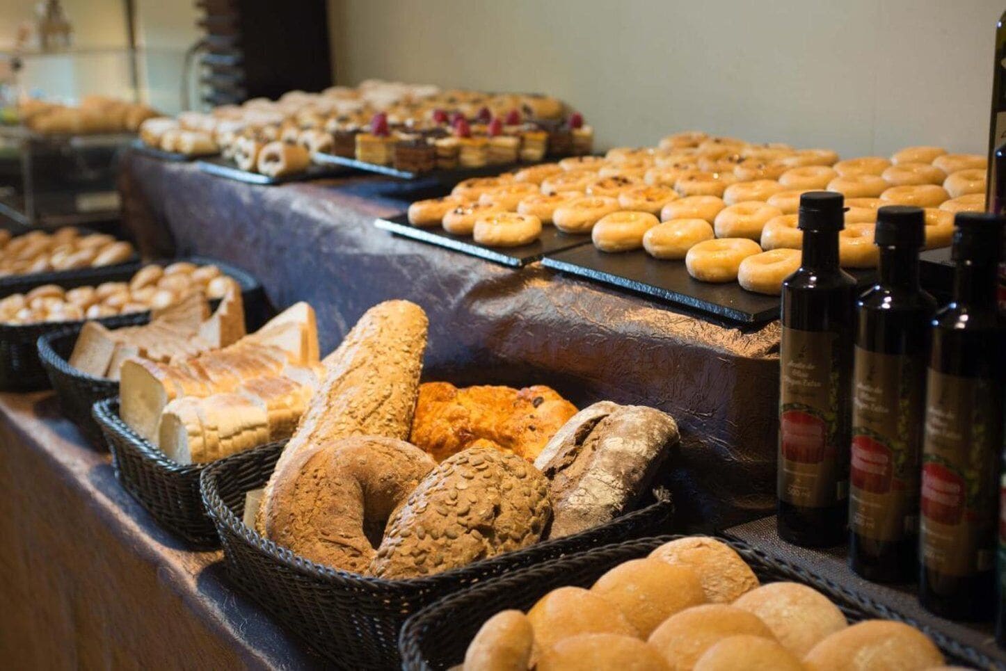 Variedad de panes y pasteles expuestos en un mostrador, con botellas de aceite de oliva visibles al fondo.