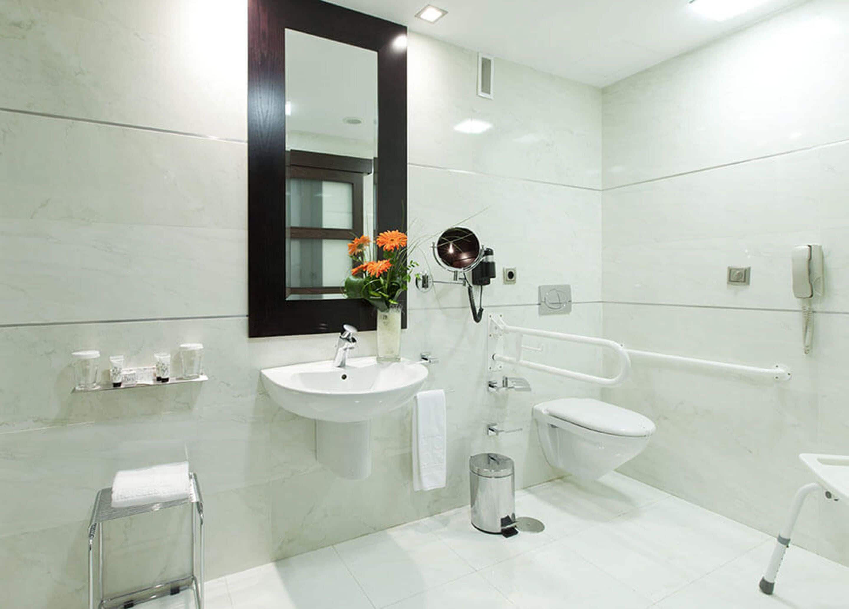 A modern bathroom with a white sink, mirror, and toilet. There is a hand dryer and a trash can.