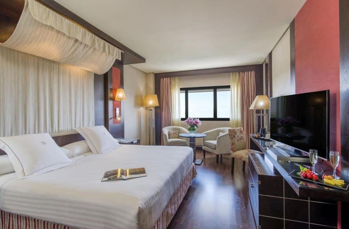 A hotel room with a large bed, a desk with a TV, a small table with chairs, and a window with curtains.