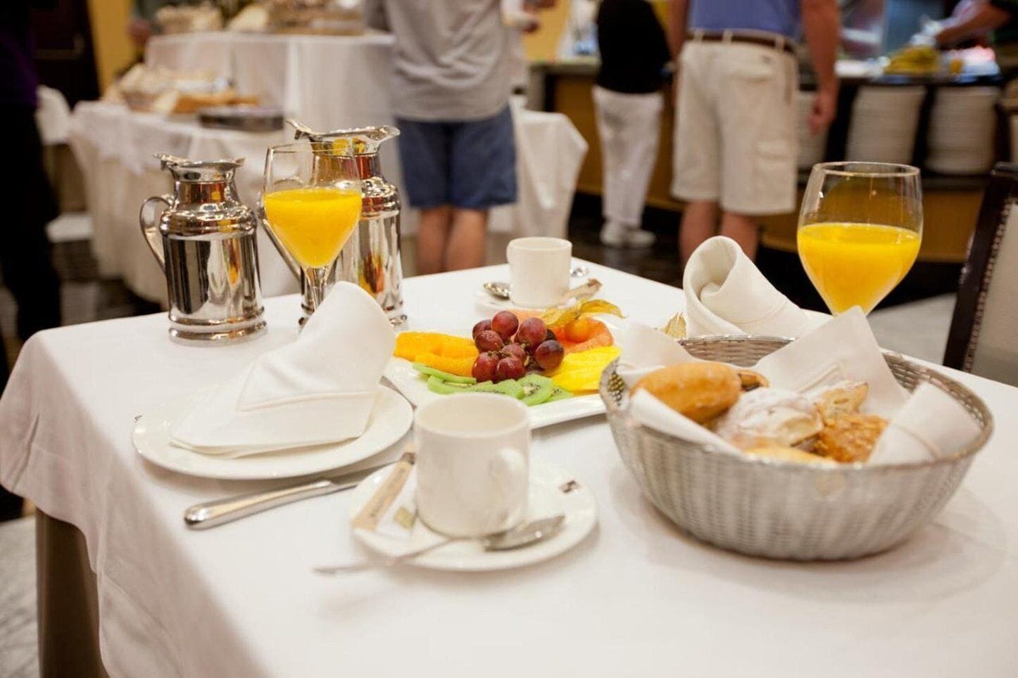 A breakfast table set with a pitcher of orange juice, a glass of orange juice, a plate of fruit, a bowl of pastries, and a cup of coffee.