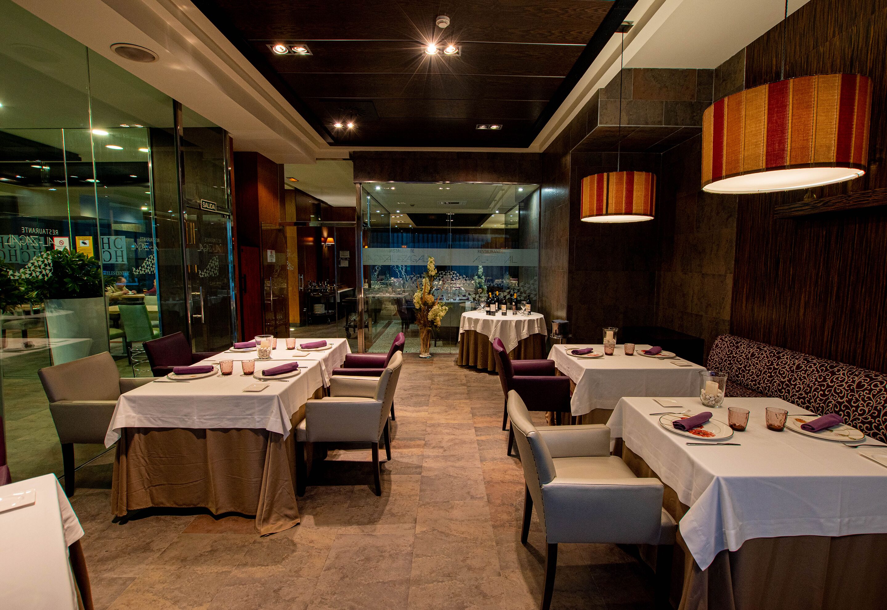 A restaurant with multiple tables and chairs, a patterned bench, and a glass wall with a view.