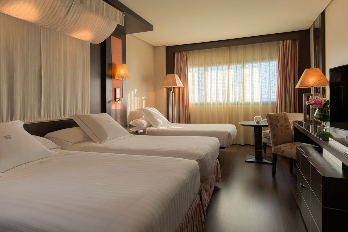 A hotel room with two beds, a desk, a chair, a TV, and a window with curtains.