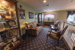 A room with a patterned carpet, a sofa, a chair, a table with a vase, and a display case with various items. - Hearthstone Inn & Suites (Cedarville)