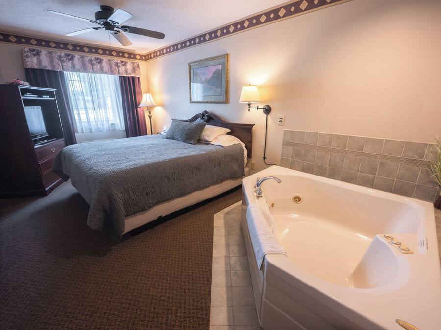 A hotel room with a bed, a bathroom with a bathtub, a ceiling fan, and a window with curtains.
