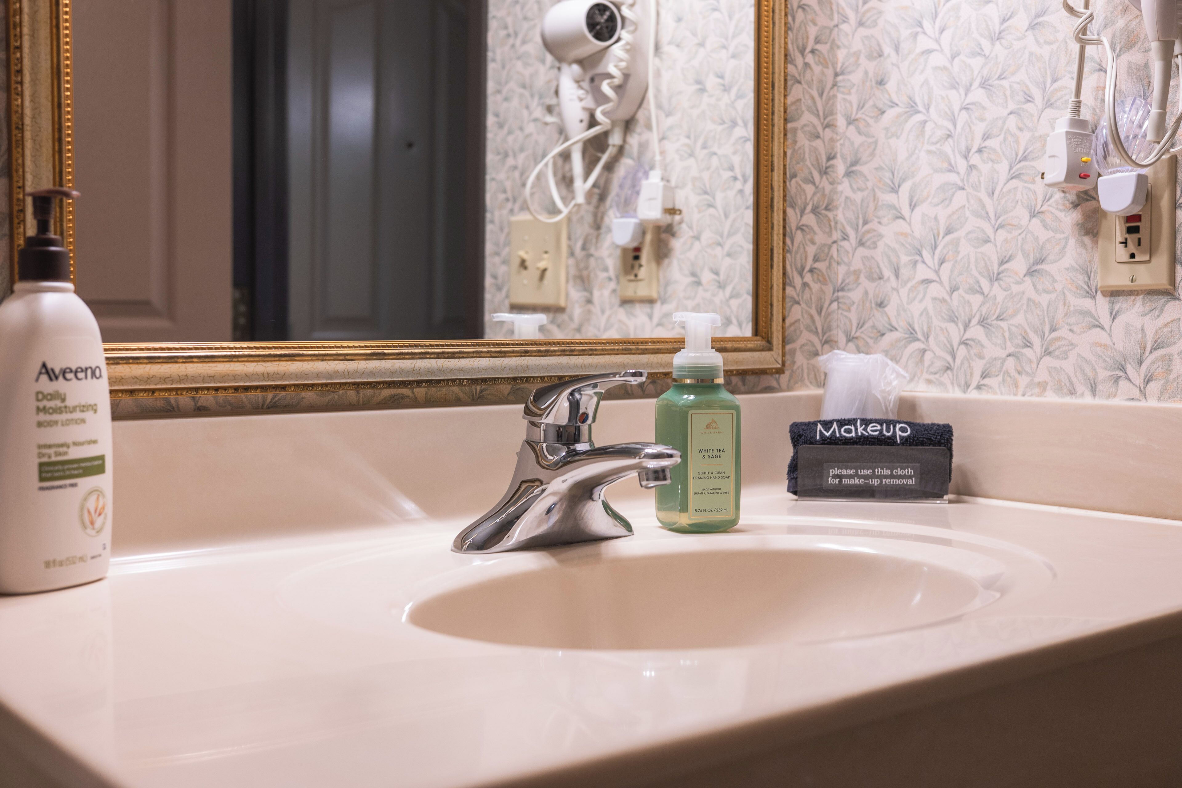 Un lavabo amb mirall, una ampolla de loció corporal Aveeno, una ampolla de sabó White Tea & Sage, i un drap per treure la maquiatge.