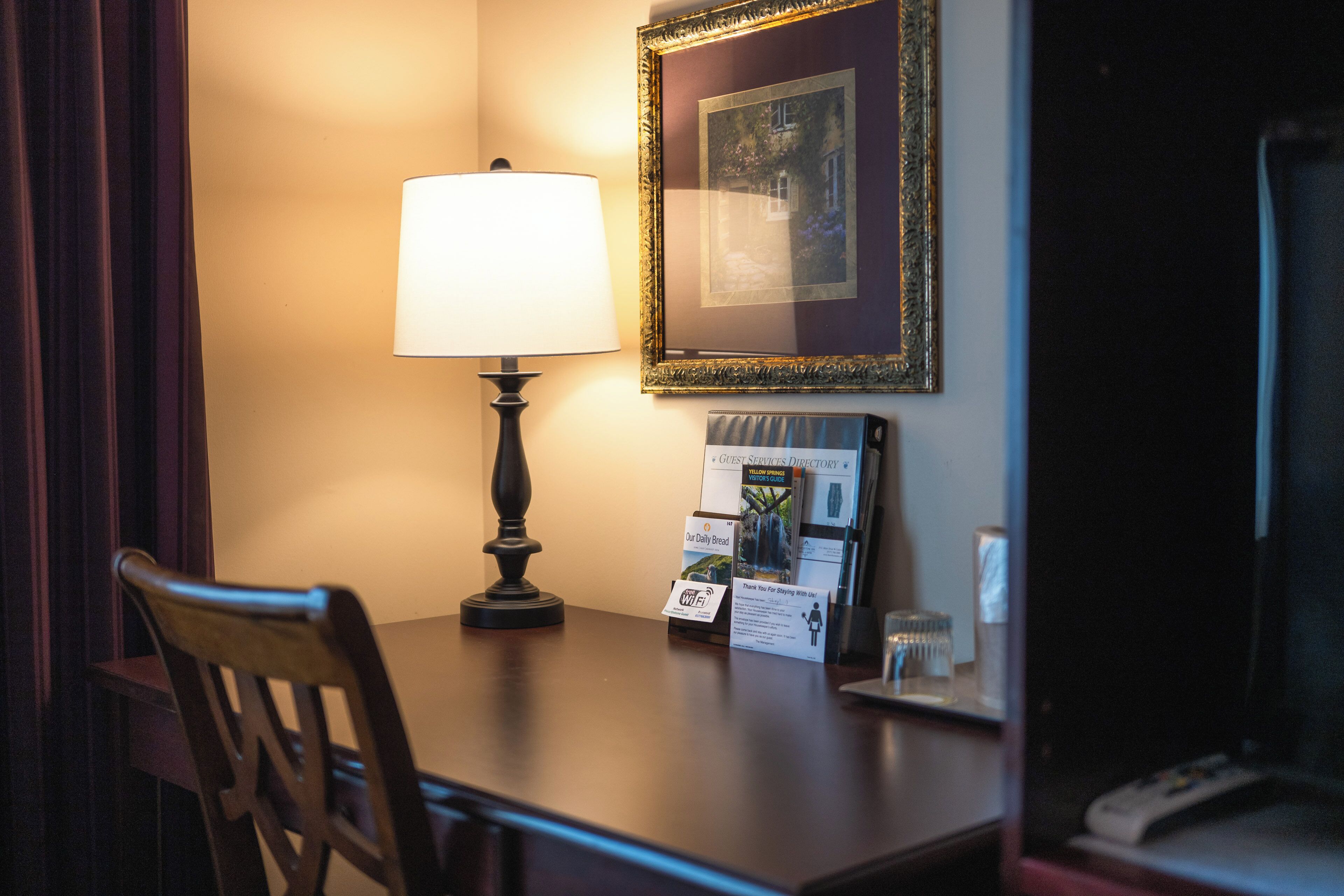 Habitación de hotel con escritorio, lámpara, un cuadro enmarco y una guía de servicios para huéspedes.