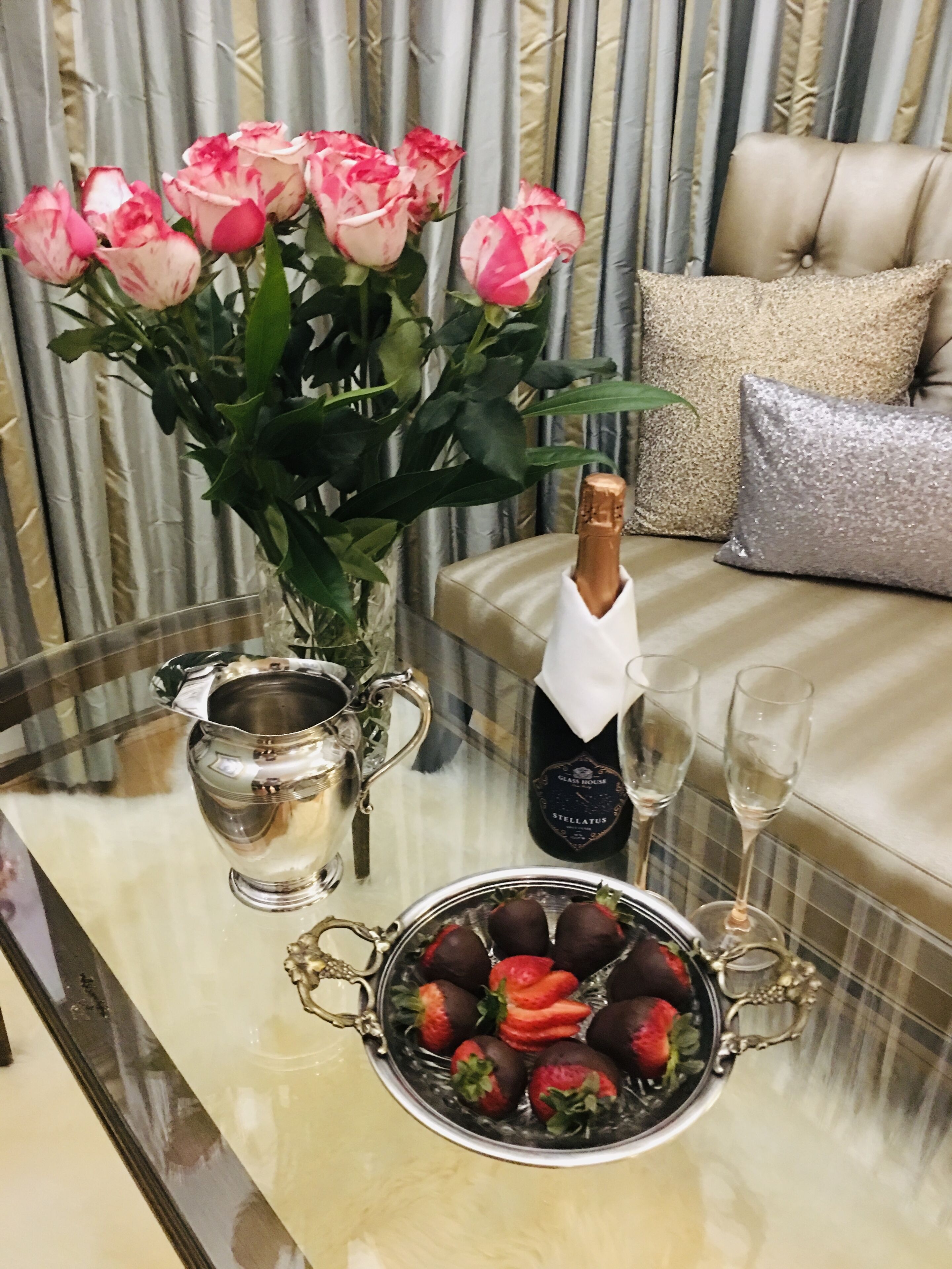 A table with a vase of pink roses, a bottle of champagne, a pitcher, and a bowl of strawberries.