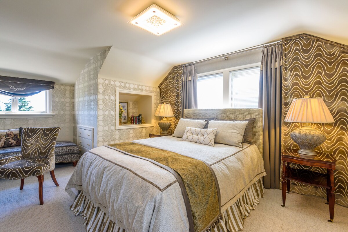 A bedroom with a large bed, a bedside table with a lamp, a chair, and a window with curtains.