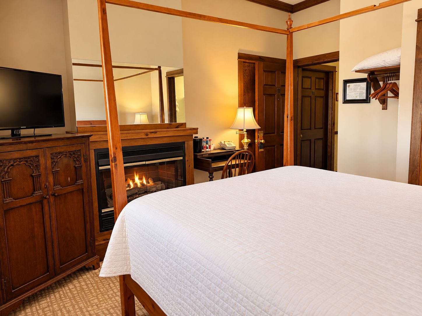 A hotel room with a four-poster bed, a fireplace, a television, and a desk with a lamp.