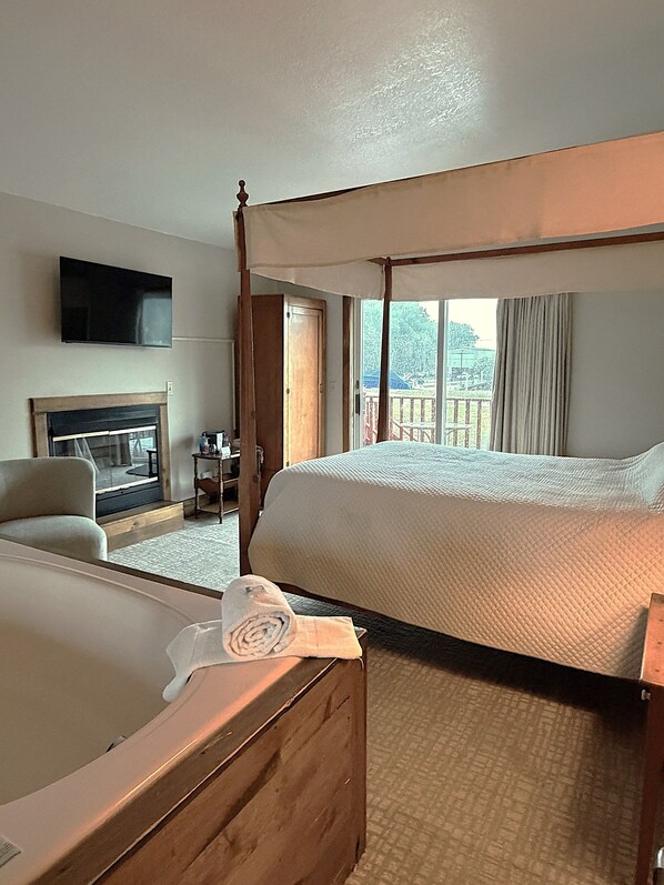 A hotel room with a four-poster bed, a flat-screen TV mounted on the wall, a fireplace, and a view of the outdoors.