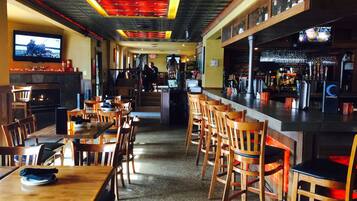 A restaurant interior with wooden tables and chairs, a bar area with a counter and stools, and a television mounted on the wall.