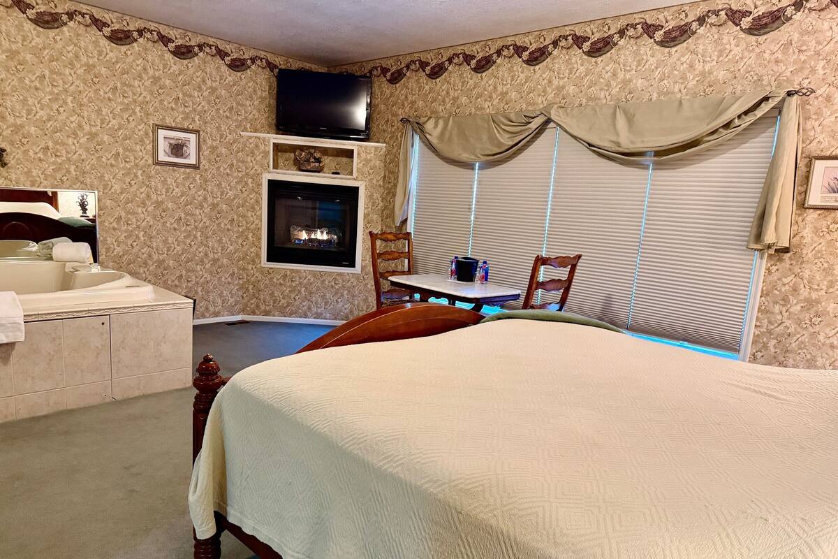 A bedroom with a large bed, a vanity with a mirror, a fireplace, and a dining area with a table and chairs.