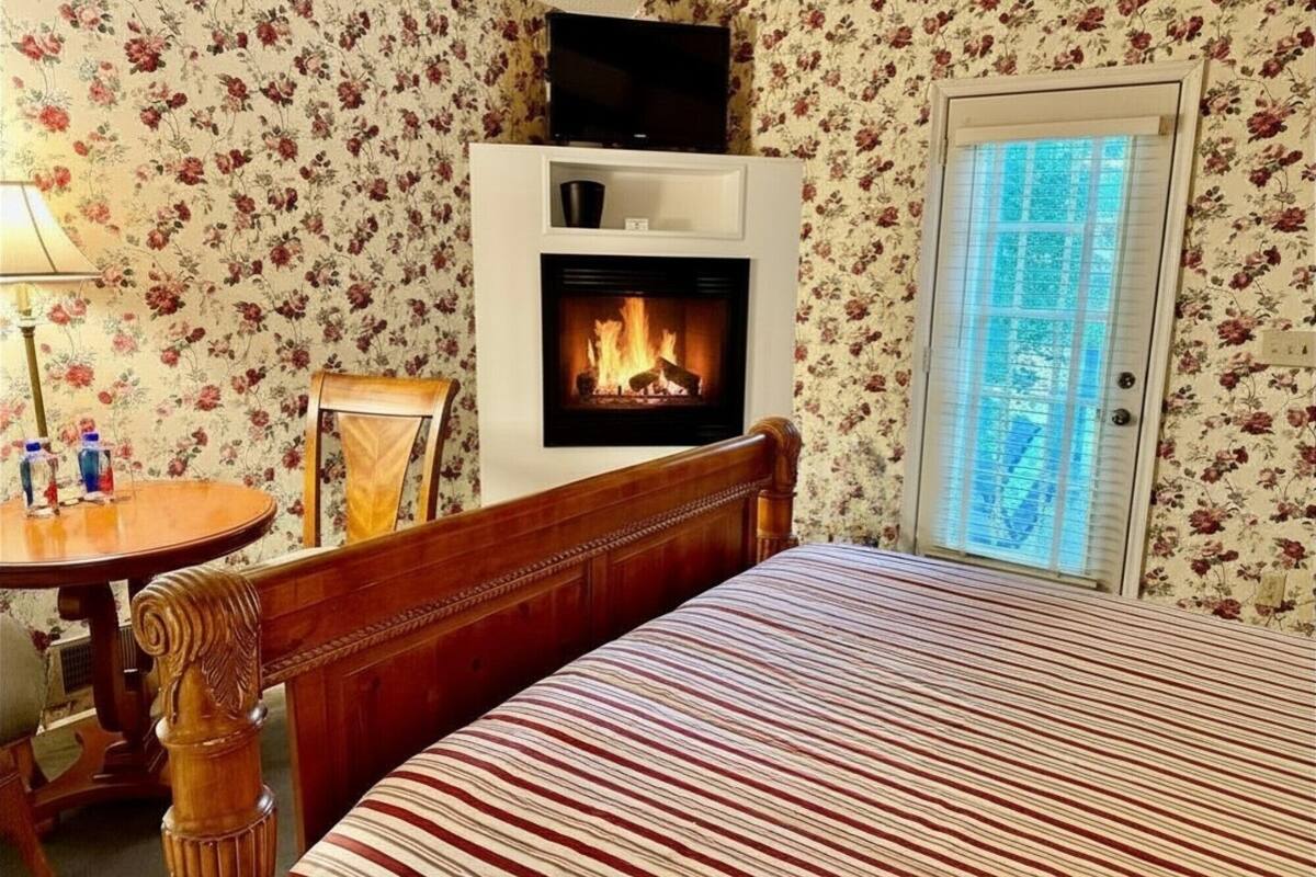 A bedroom with a bed, a fireplace, a television, a chair, a table, and a door with a window.