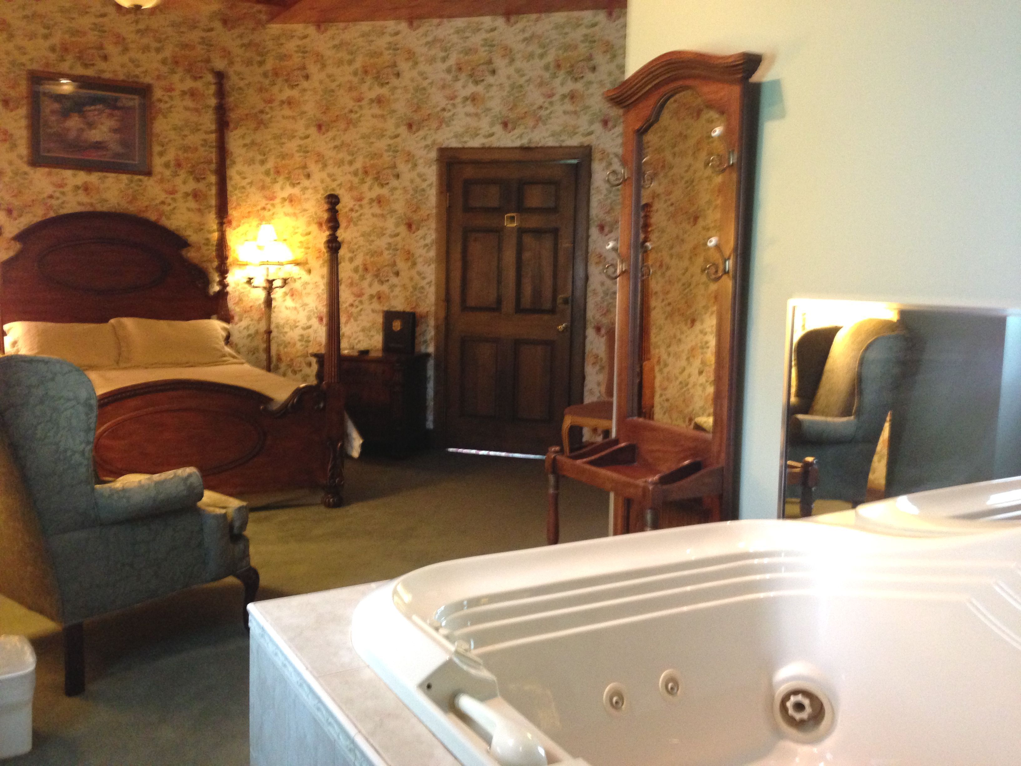 A bedroom with a bed, a chair, a wooden wardrobe, a mirror, and a bathtub.