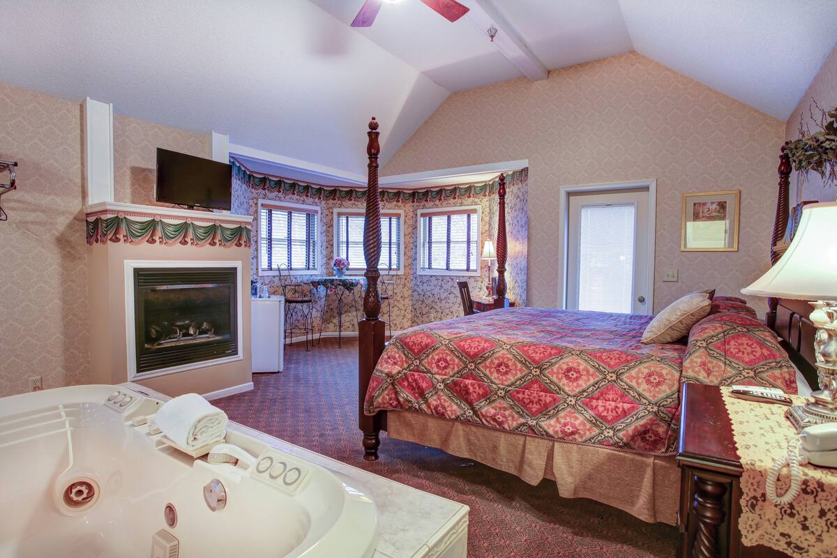 A bedroom with a four-poster bed, a fireplace, a large window, and a bathroom with a bathtub.