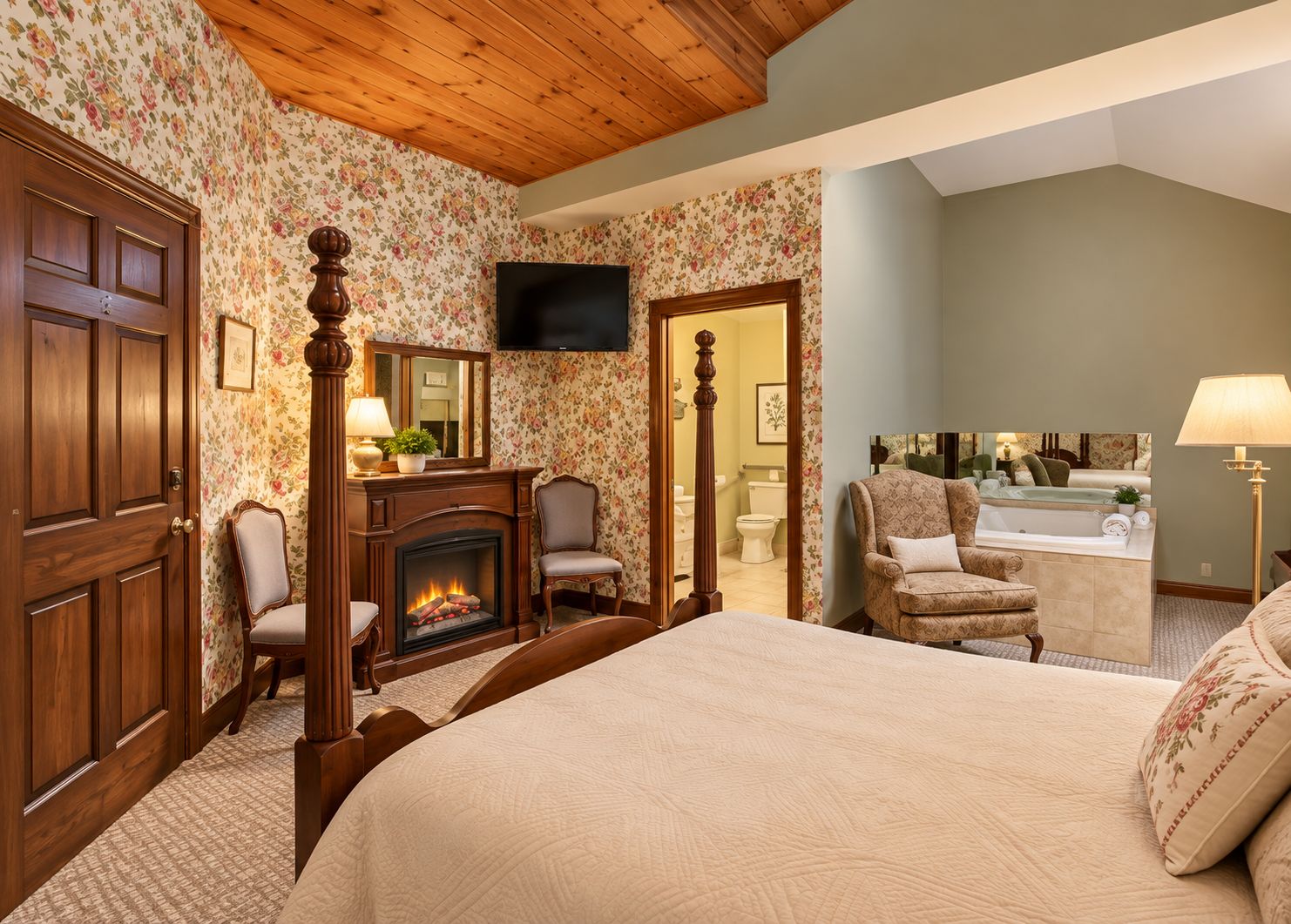 A hotel room with a fireplace, a bed, a sitting area with a chair, a television, and a bathroom visible through an open door.