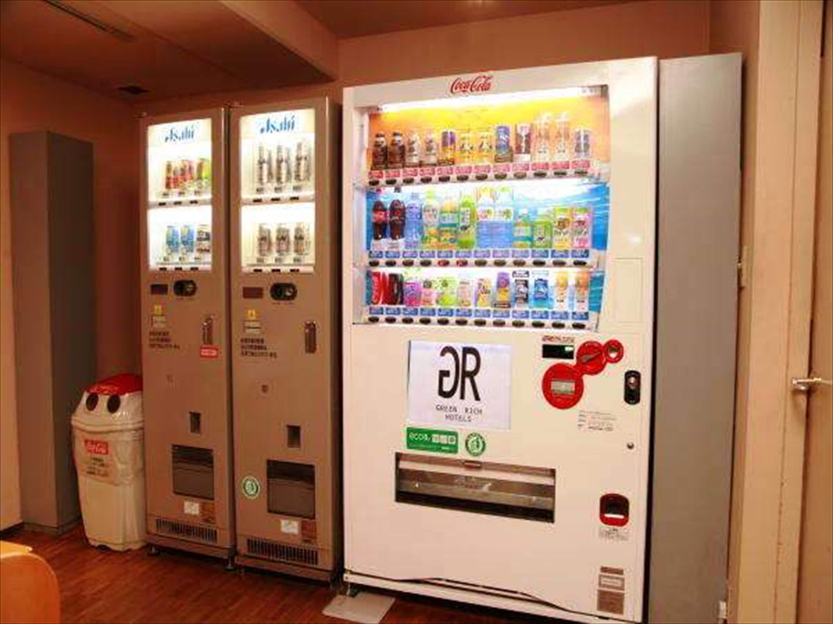 A row of vending machines with Coca-Cola and other beverages, and a recycling bin.