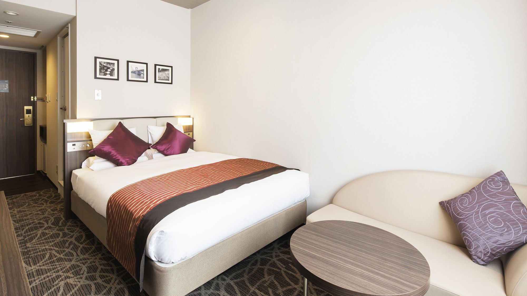A hotel room with a bed, a sofa, a small table, and three framed pictures on the wall.