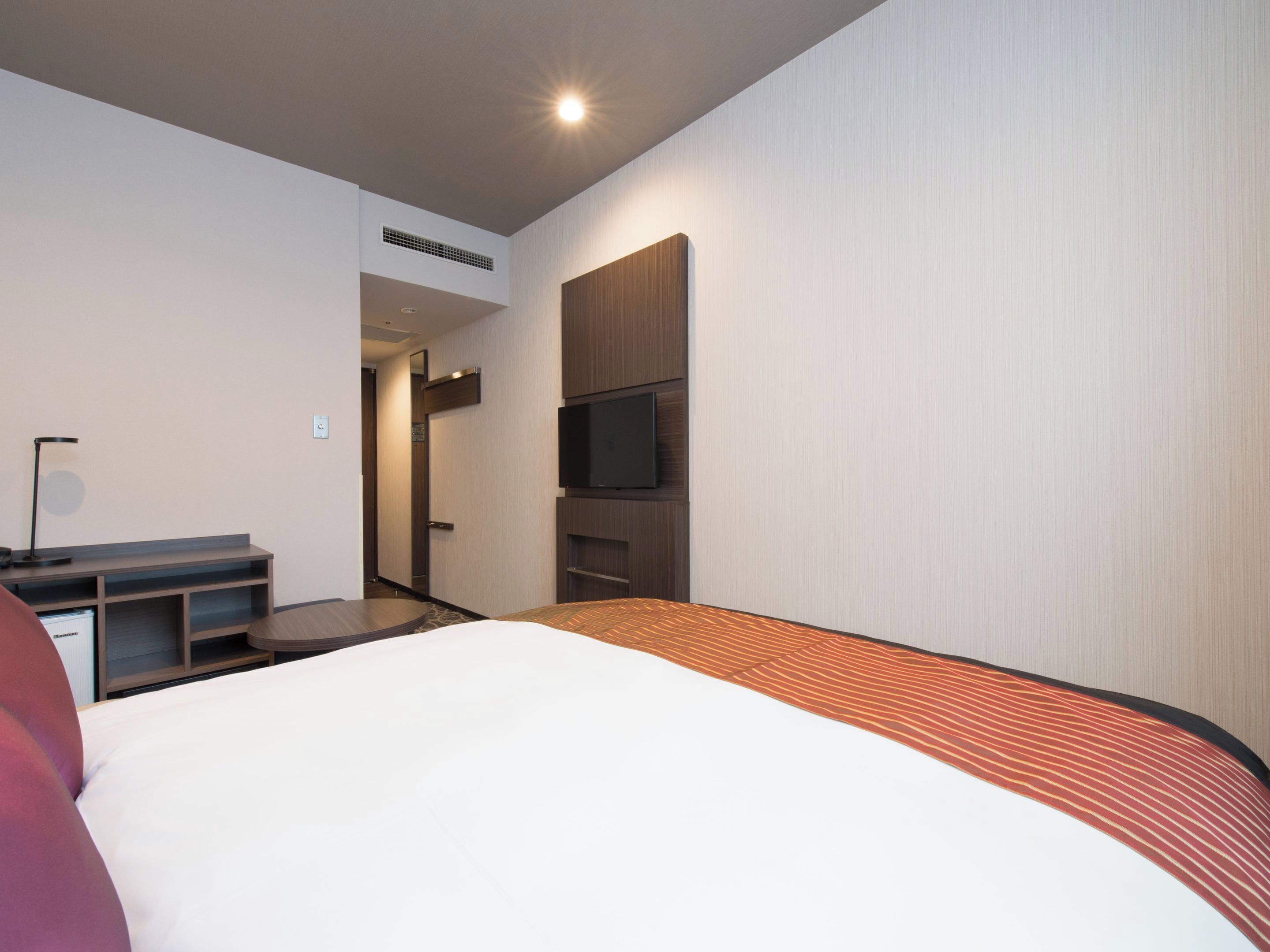 A modern hotel room with a bed, a TV mounted on the wall, a desk with a lamp, and a door leading to another room.
