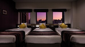 A hotel room with three beds, each with a striped bedspread and a pillow. The room has large windows with a cityscape view at sunset. - HOTEL MYSTAYS Ueno East (Tokyo)