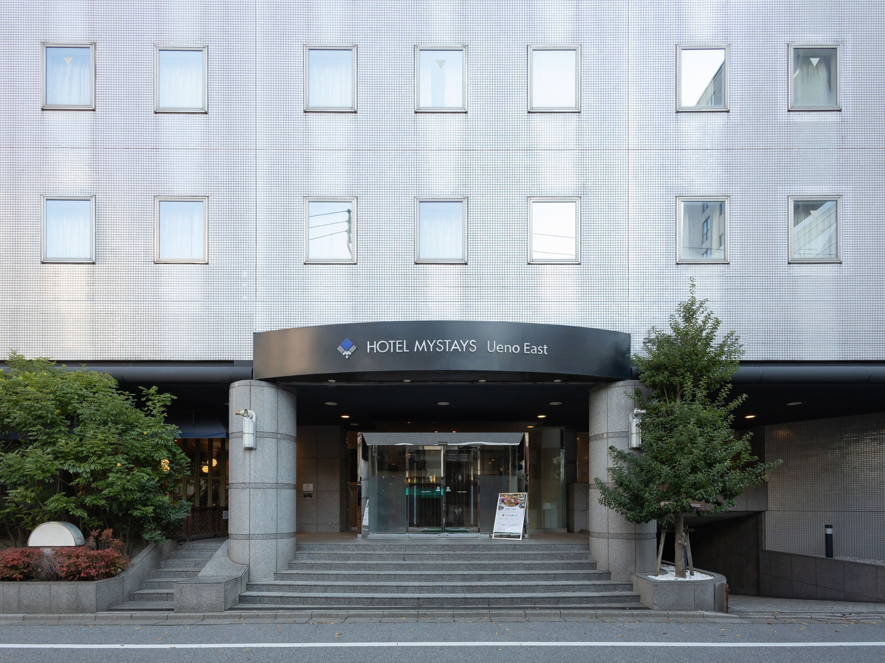 Une entrée d’hôtel moderne avec une façade carrelée, une enseigne indiquant « HÔTEL MYSTAYS Ueno Est » et un escalier menant à l’entrée.