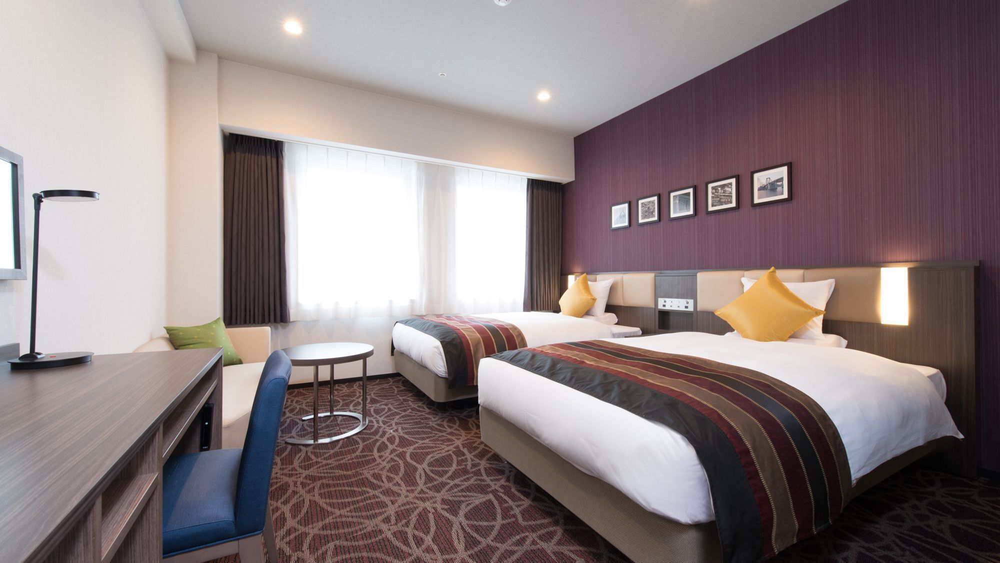 A hotel room with two beds, a desk, a chair, a small table, and a large window with curtains.