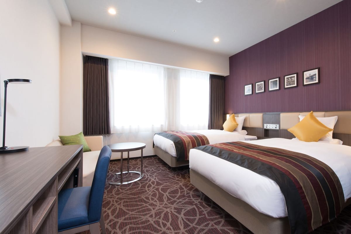 Une chambre d’hôtel avec deux lits, un bureau, une chaise, une petite table et une grande fenêtre avec des rideaux.