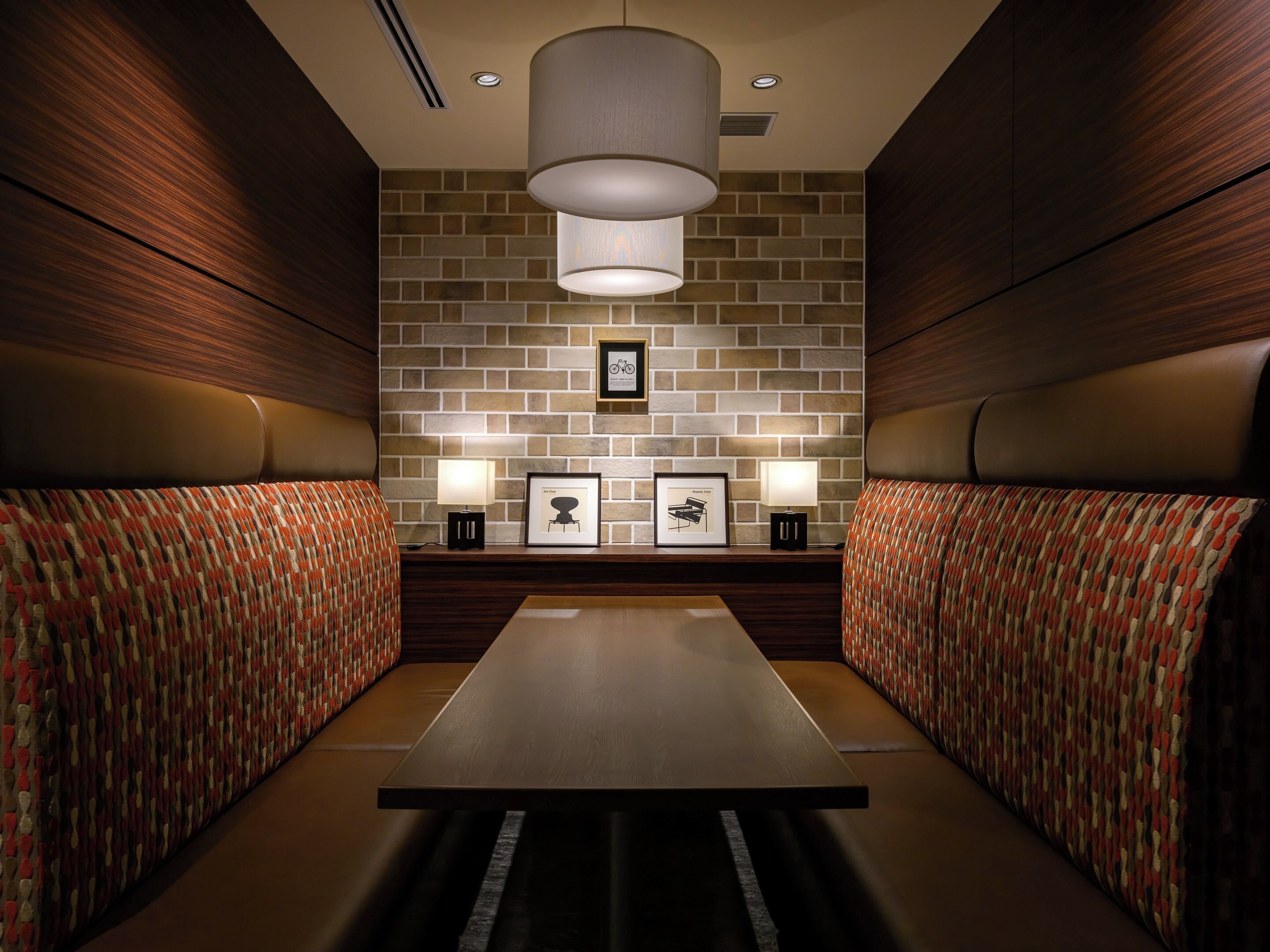 A hotel room with a brick-patterned wall, two cushioned seating areas, a central table, and two wall-mounted lamps.