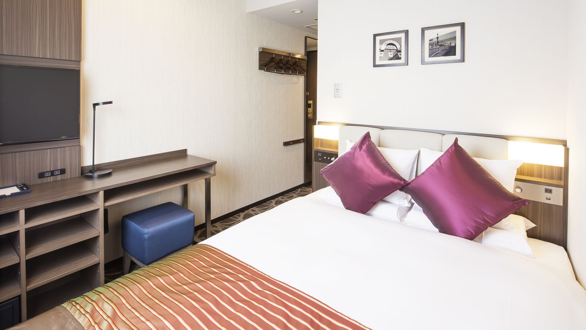 A hotel room with a bed, a desk, a TV, and two framed pictures on the wall.