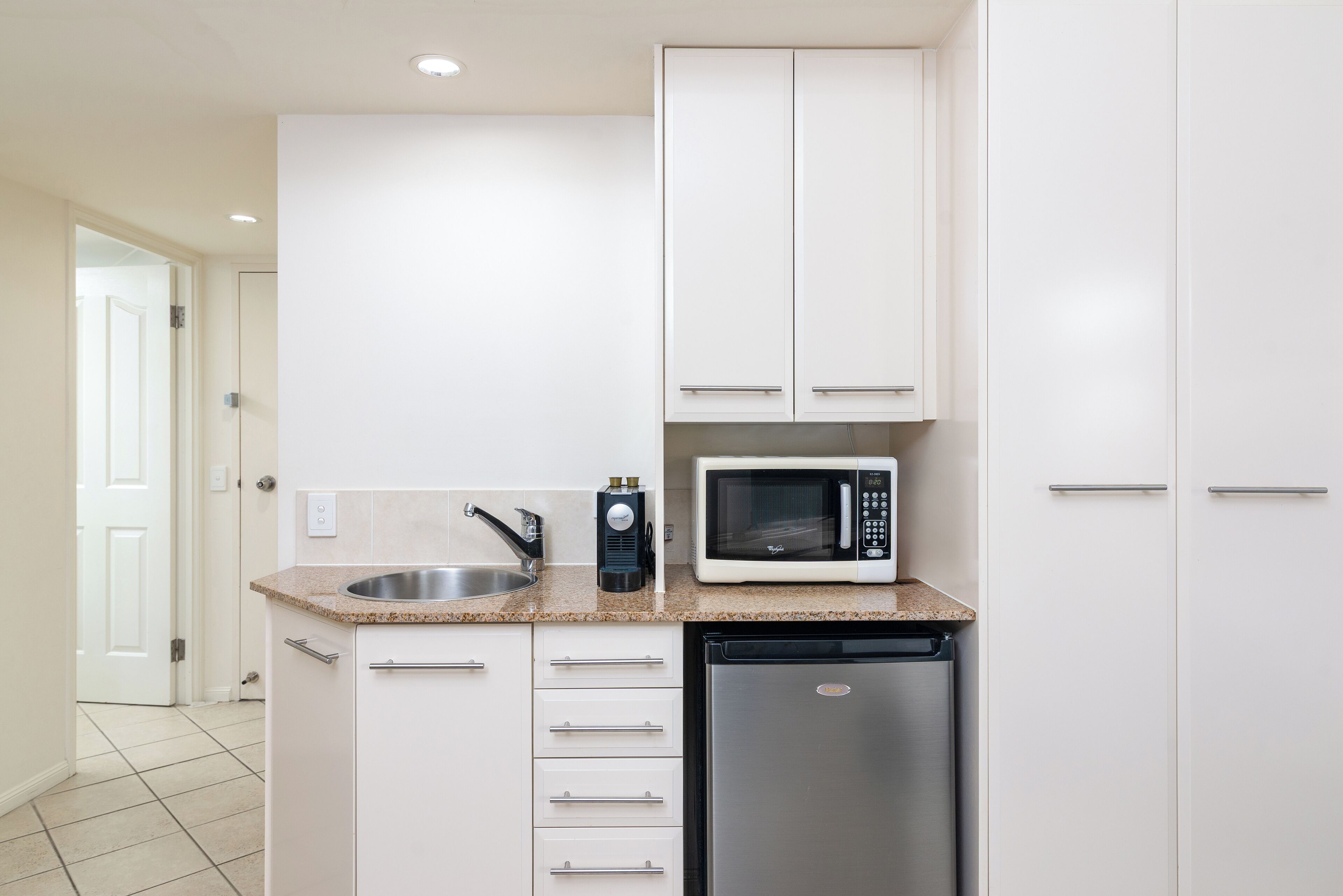Una cocina compacta con muebles blancos, un lavamanos de acero inoxidable, un microondas y un lavavajillas.