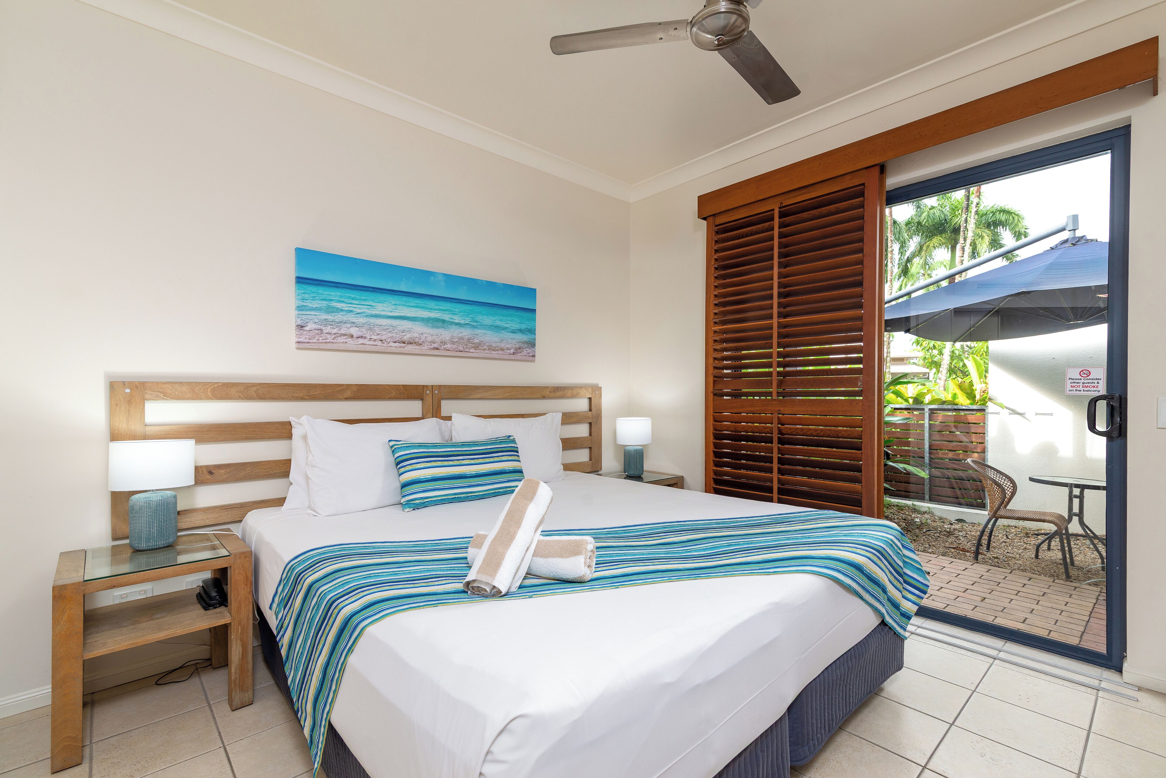 A hotel room with a bed, nightstands, a ceiling fan, a beach painting, and a view of the outdoors.