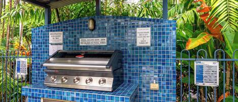 A blue-tiled BBQ area with a stainless steel grill, surrounded by lush tropical plants.
