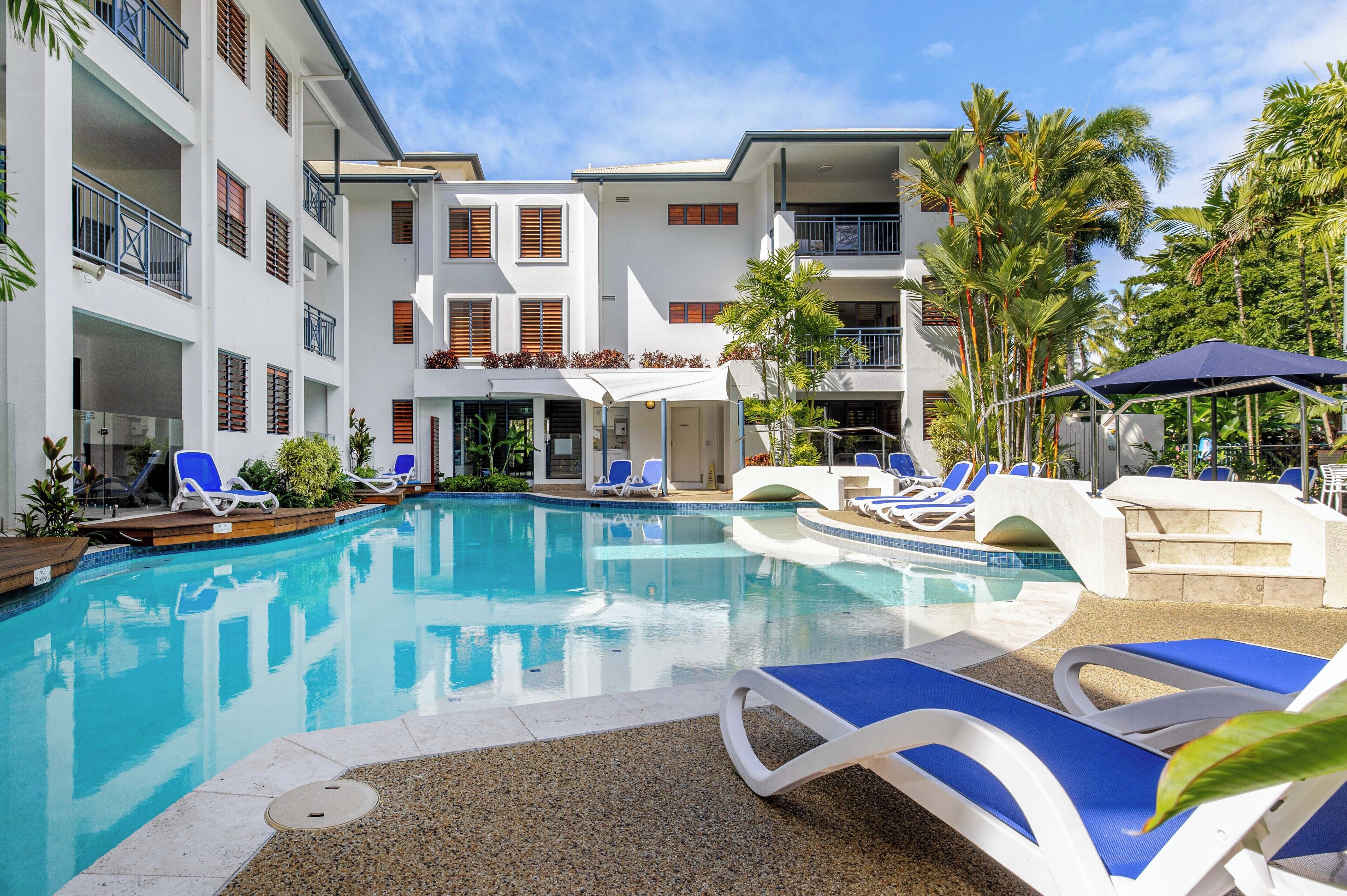 Une piscine avec des chaises longues, des palmiers et un immeuble à plusieurs étages doté de balcons.