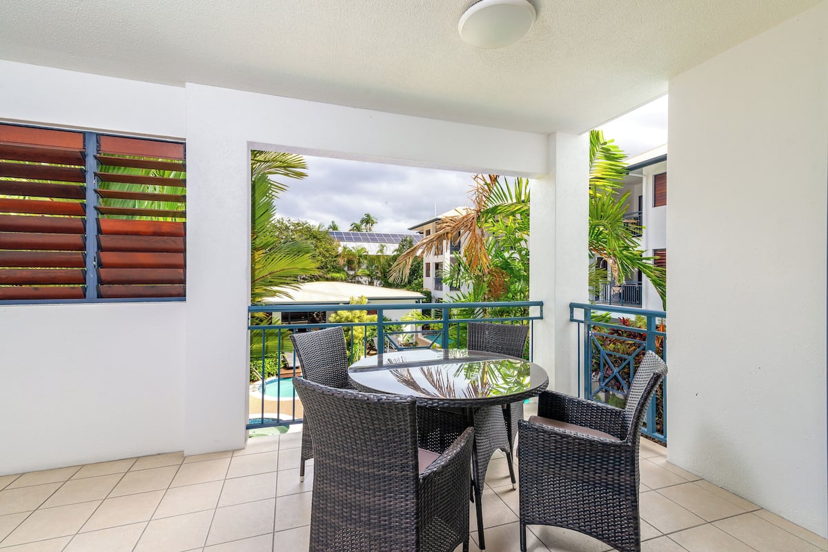 Ein Balkon mit rundem Tisch und Korbstühlen, der einen Pool und üppiges Grün blickt.