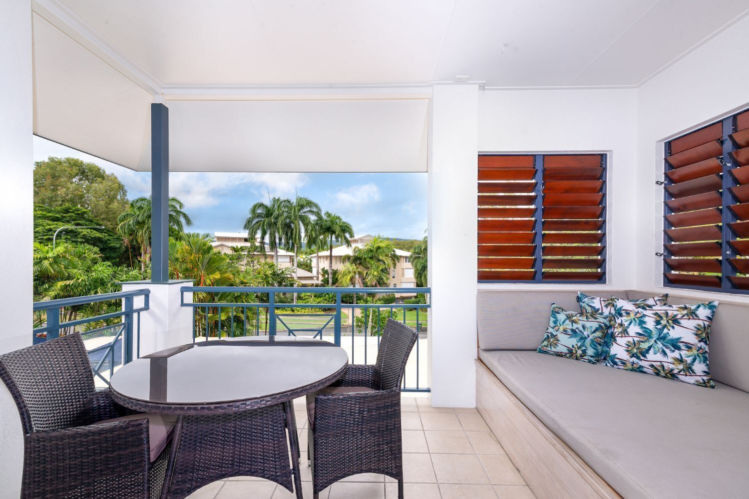 A balcony with a table and chairs, a sofa with patterned cushions, and a view of palm trees and buildings.