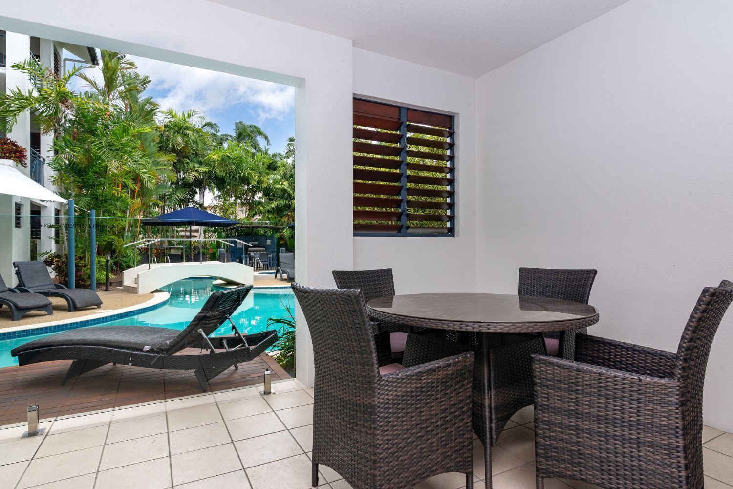 A poolside area with a round table and chairs, a pool, and a view of a building with a covered seating area.