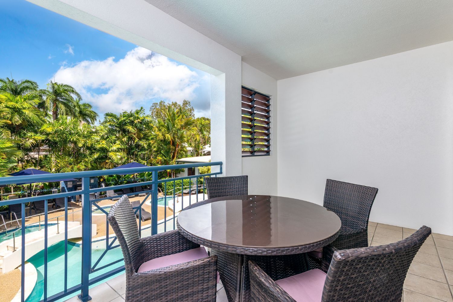 A balcony with a round table and wicker chairs, overlooking a pool and palm trees.