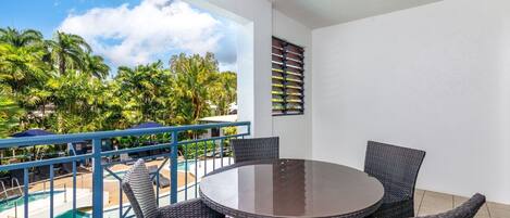 A balcony with a round table and wicker chairs, overlooking a pool and palm trees.