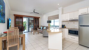 A modern kitchen with white cabinets, a stainless steel refrigerator, and a built-in oven. There is a dining area with a wooden table and chairs, and a living room with a sofa and a coffee table. - Meridian Port Douglas - Adults only (Port Douglas)