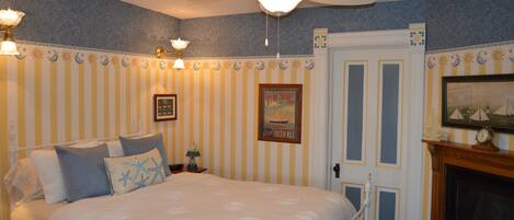 A bedroom with a bed, a ceiling fan, a nightstand, a lamp, a framed picture, and a fireplace.