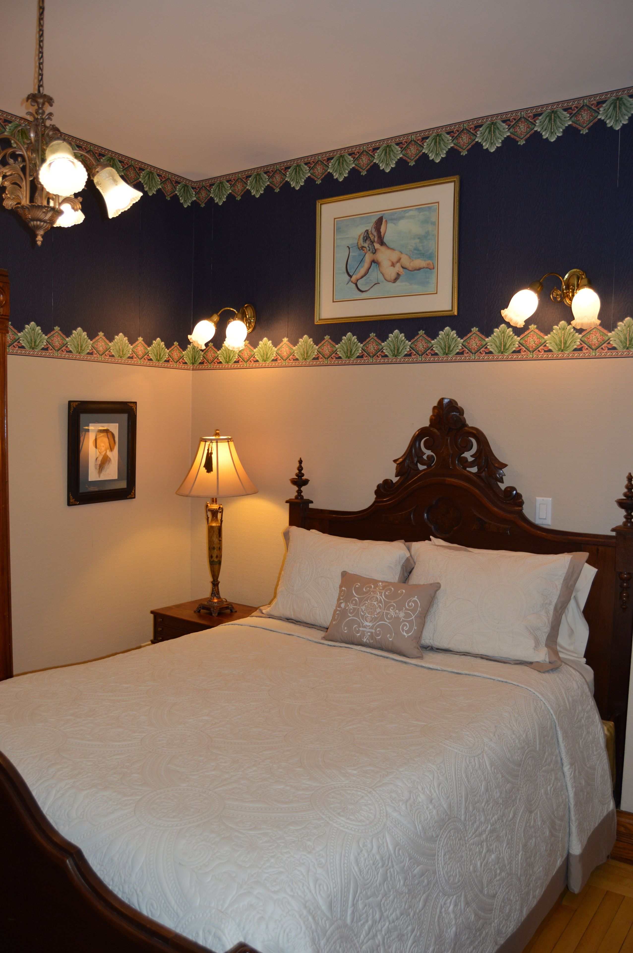 Un lit bien fait, avec une couette blanche et plusieurs oreillers, une lampe de chevet, un tableau encadré au mur et un luminaire décoratif au plafond.