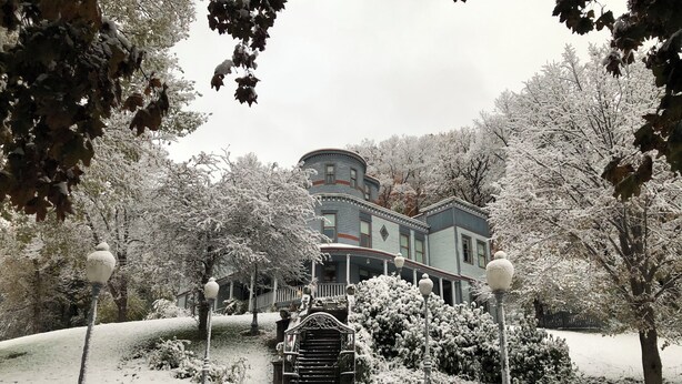 A snow-covered pathway leading to a historic house with a porch and a staircase.