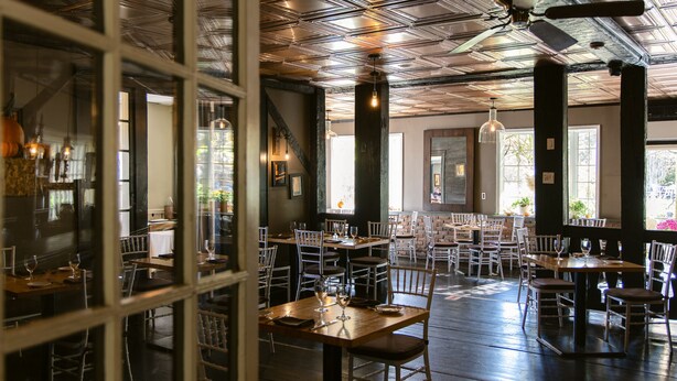 A restaurant interior with multiple dining tables and chairs, a ceiling with exposed beams, and hanging pendant lights. - The Kittle House Inn (Chappaqua)