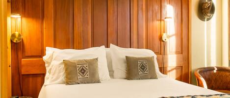 A hotel room with a wooden paneled wall, a bed with white linens, two decorative pillows, a small round table, and a chair.